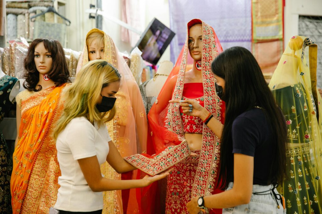 Two women browse traditional Indian sarees in a market, both wearing face masks for safety.